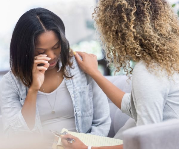 Caring therapist places her hand on a young woman's shoulder as the woman cries during a therapy session.